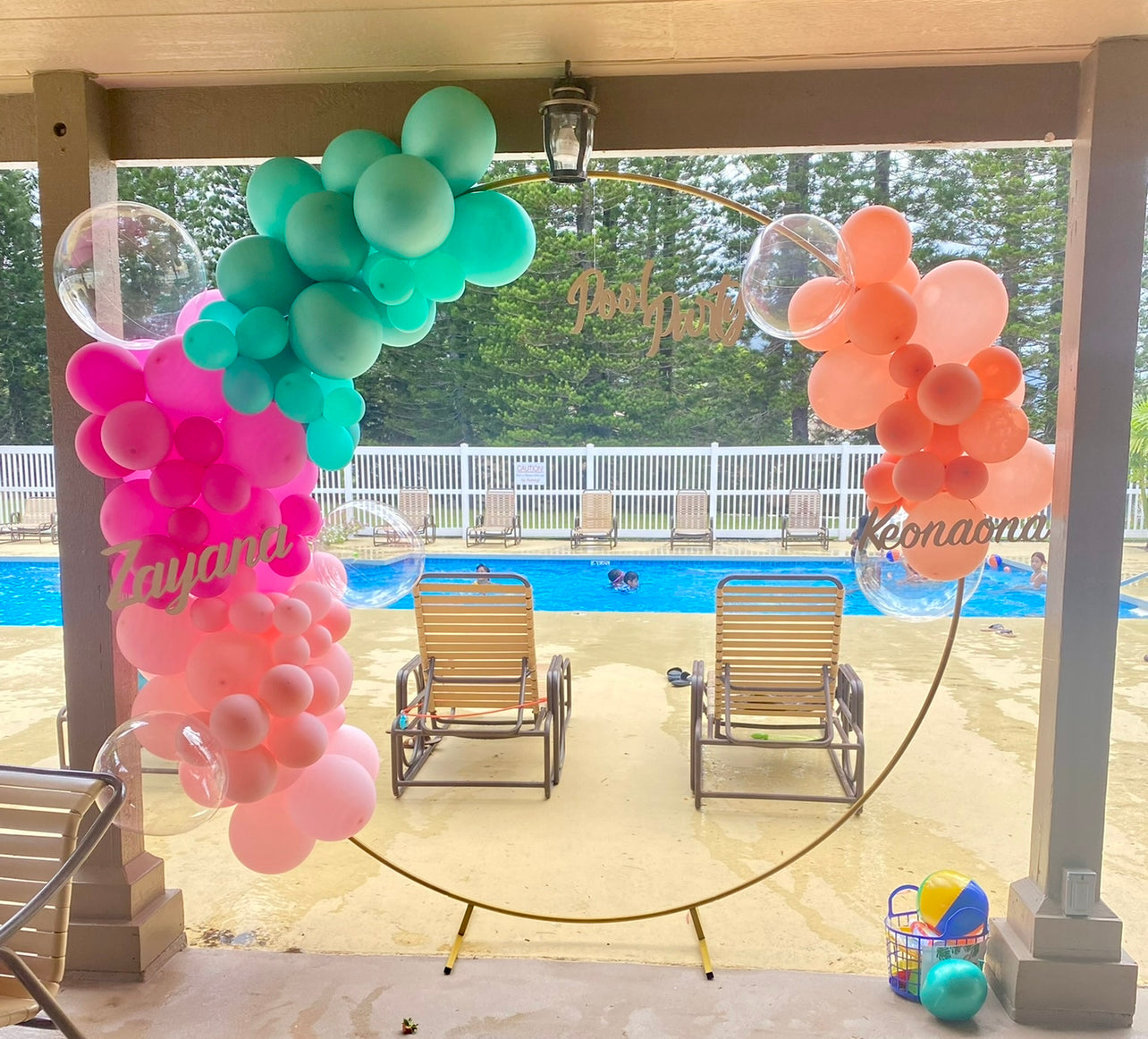 Round balloon arch stand is adorned with clusters of pool party themed color balloons. Wooden name cut outs hang from and propped onto the arch